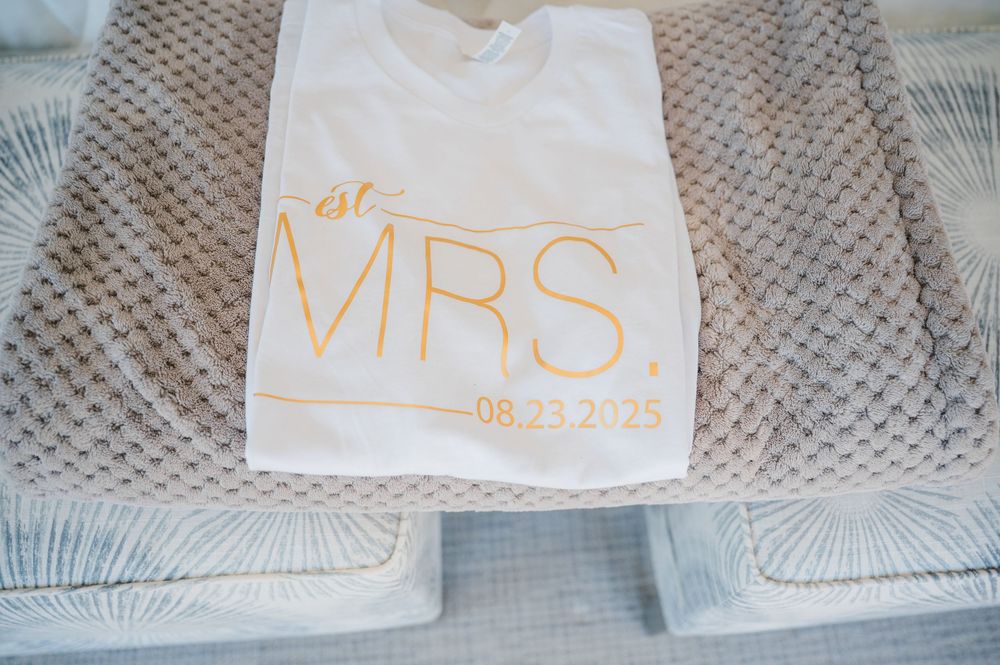 A white t-shirt with gold 'MRS' text and date embroidery lies on a textured gray surface.