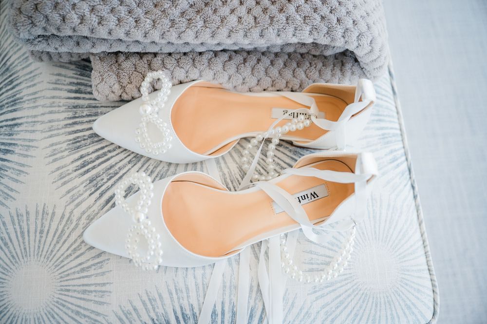 Delicate white bridal shoes with crystal embellishments displayed on a soft gray surface.