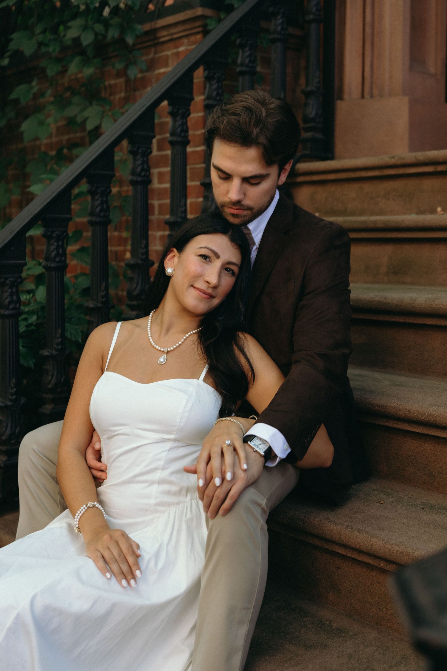 A couple in wedding attire poses romantically on brownstone steps with ivy in the background.
