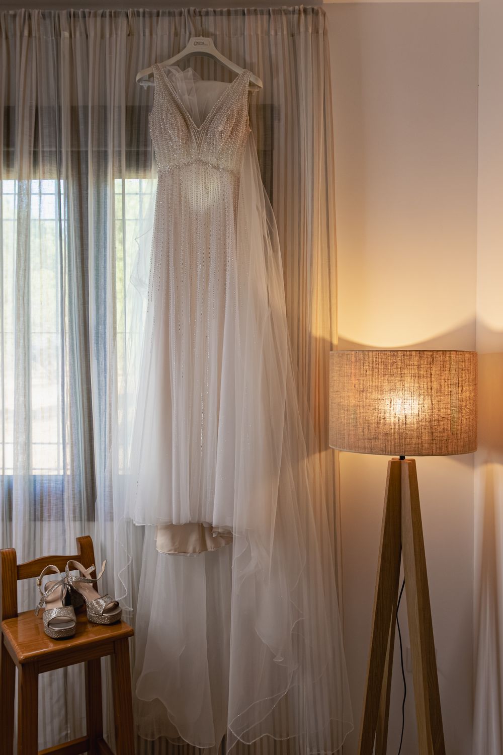 Elegant white curtains and wedding dress hanging by window with soft natural lighting and table lamp.
