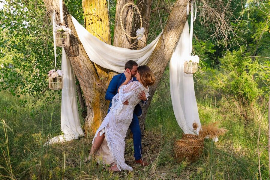 Couple shares romantic moments under draped white fabric between trees in an outdoor garden setting.