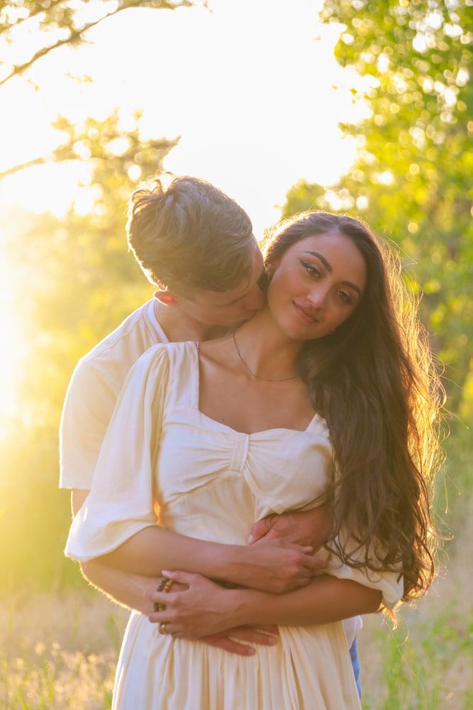 A romantic couple embraces in a sunlit meadow wearing white clothing during golden hour.