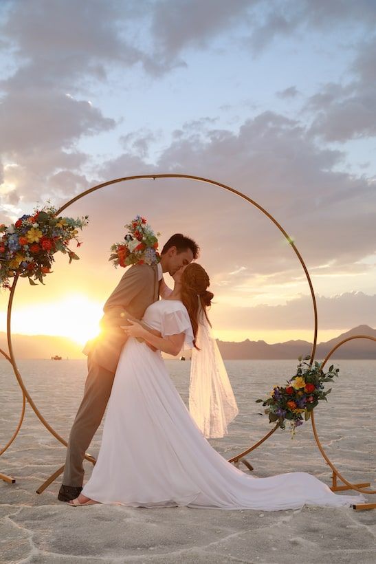 A romantic beach wedding photo series at sunset with a circular arch and floral decorations by the ocean.