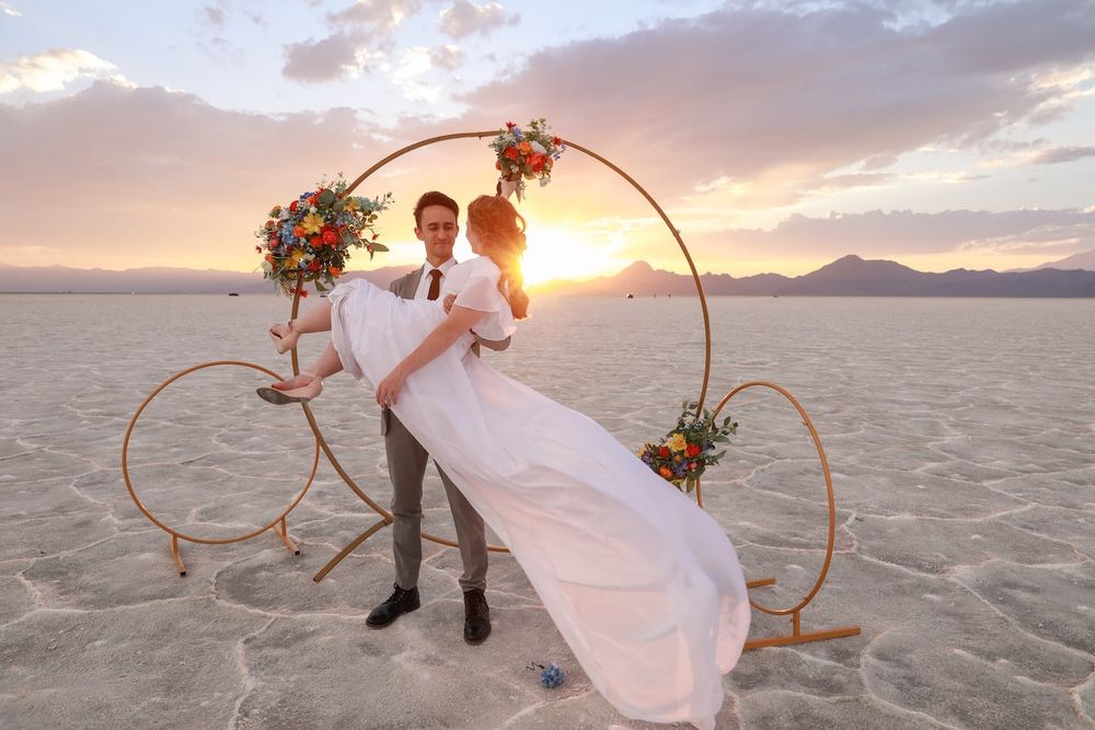 Newlyweds pose together on a salt flat at sunset with dramatic clouds and mountains in the background.