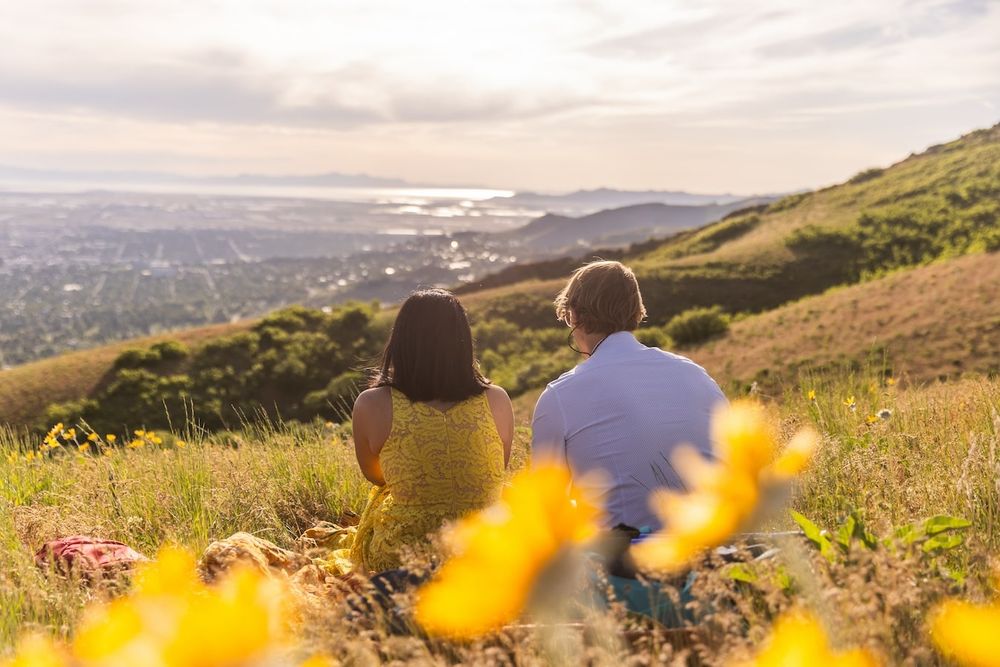 Two people sitting on grassy hillside overlooking coastal city view at sunset with yellow wildflowers in foreground.