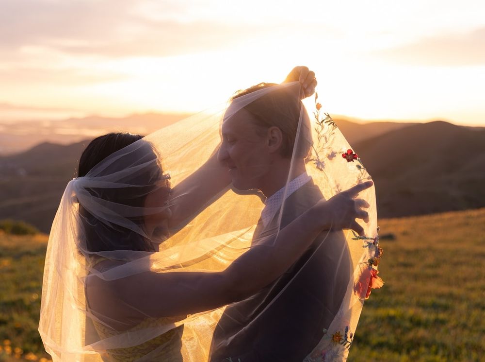 A romantic couple in wedding attire shares an intimate moment at sunset in a mountainous landscape.