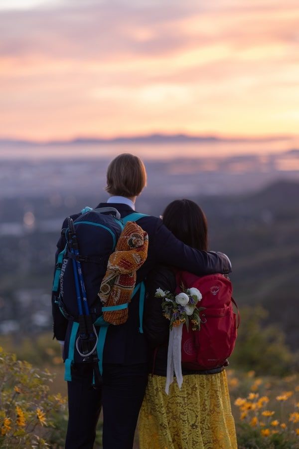 Two hikers with backpacks adorned with stuffed animals watch the sunset over mountains during golden hour.