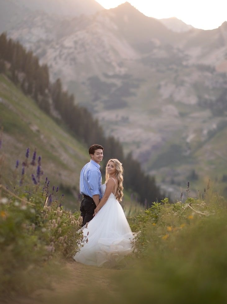 A couple stands together against a scenic mountain backdrop with purple wildflowers in the foreground.