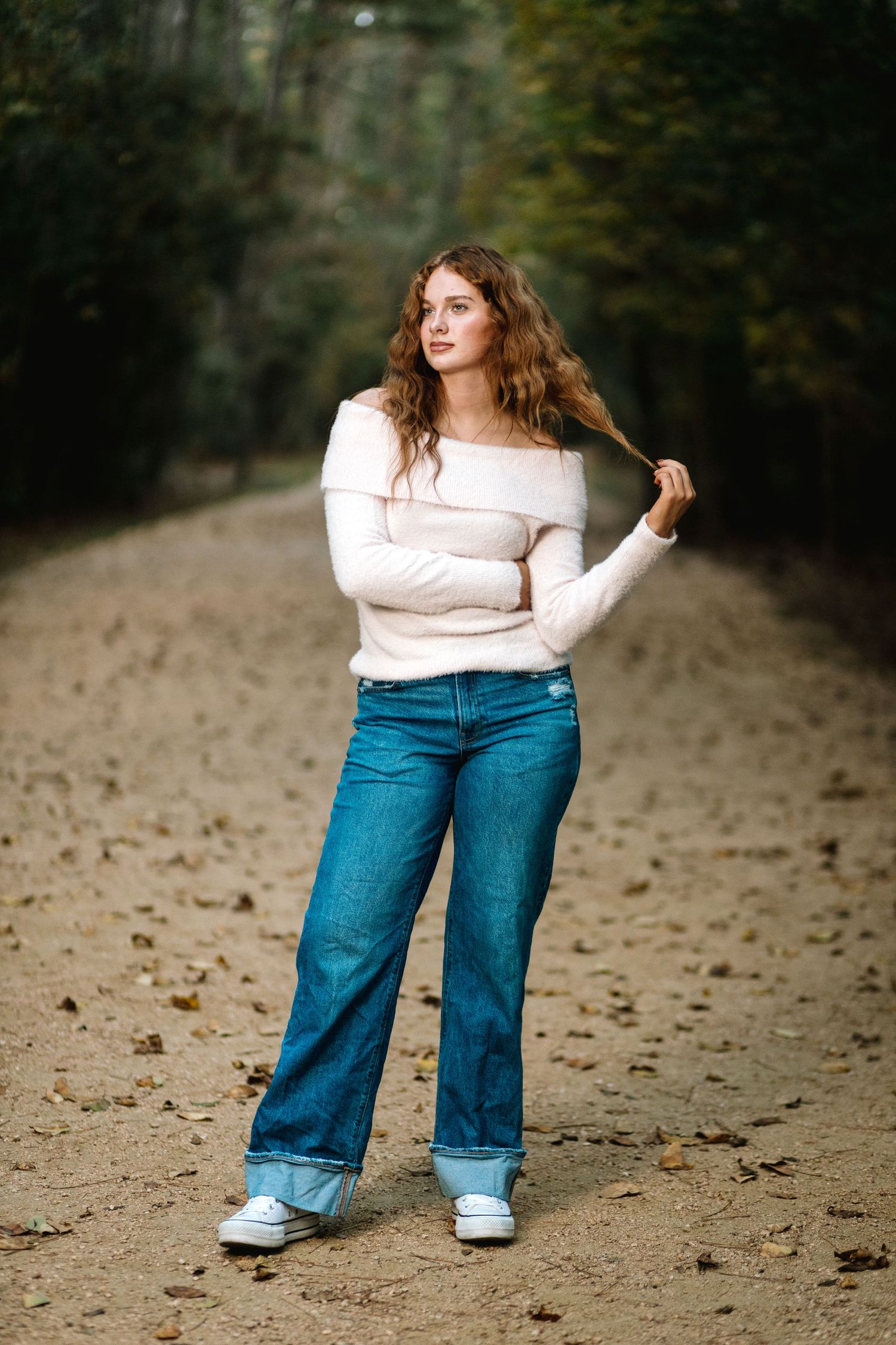 A person in a cream sweater and wide-leg blue jeans poses playfully on a dirt path surrounded by trees.