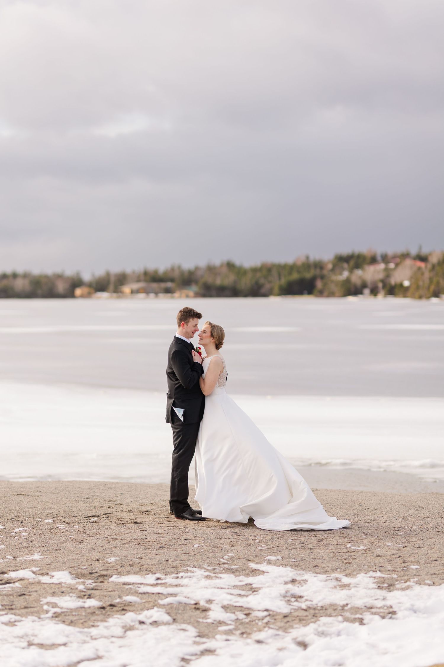 A romantic winter wedding couple embraces on a snowy beach with a lake view in the background.