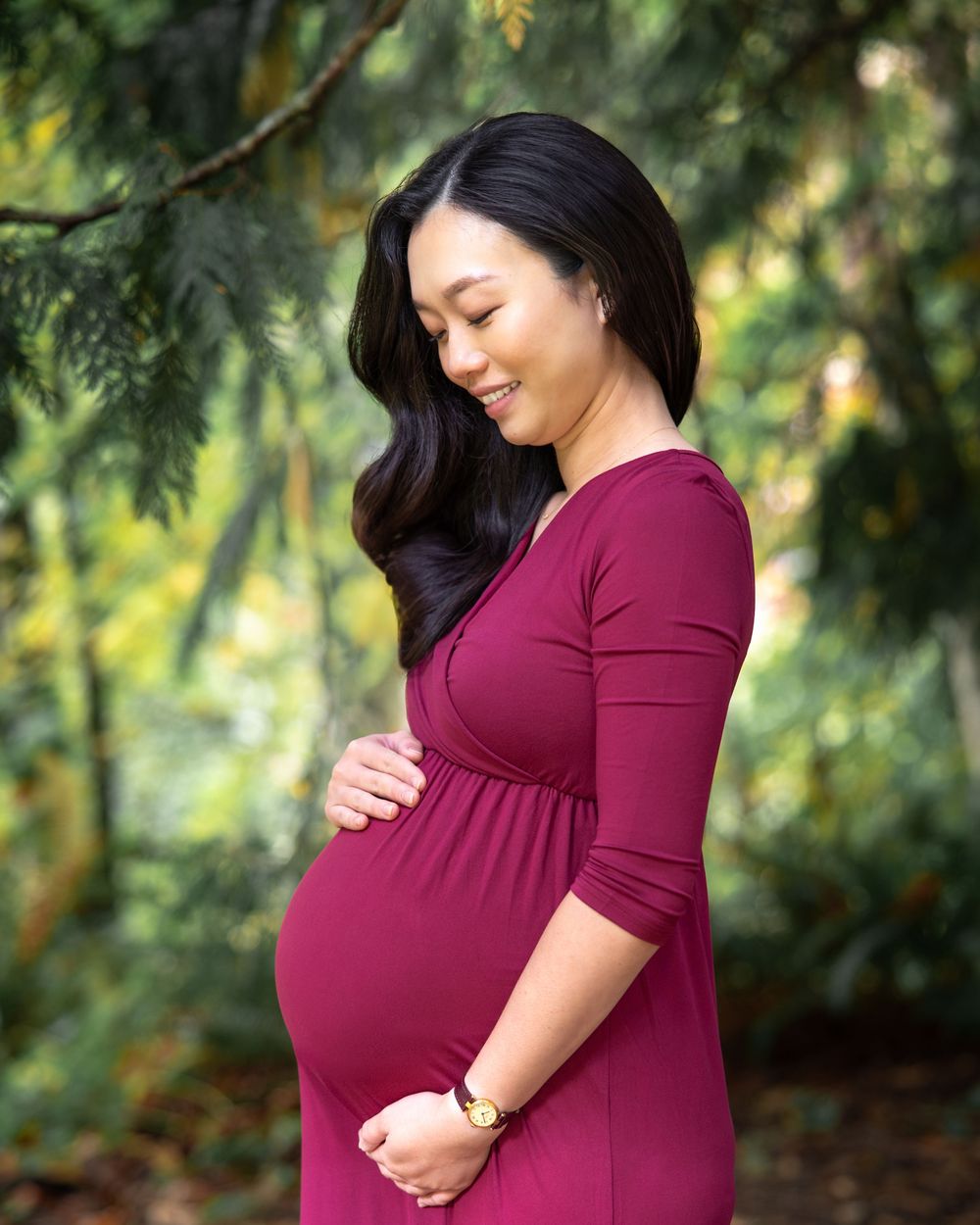 A person in a burgundy dress poses outdoors for a maternity photoshoot in a natural setting with trees.