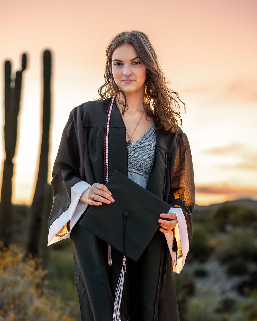 A graduate in black cap and gown holds a diploma and beverage against a sunset backdrop.