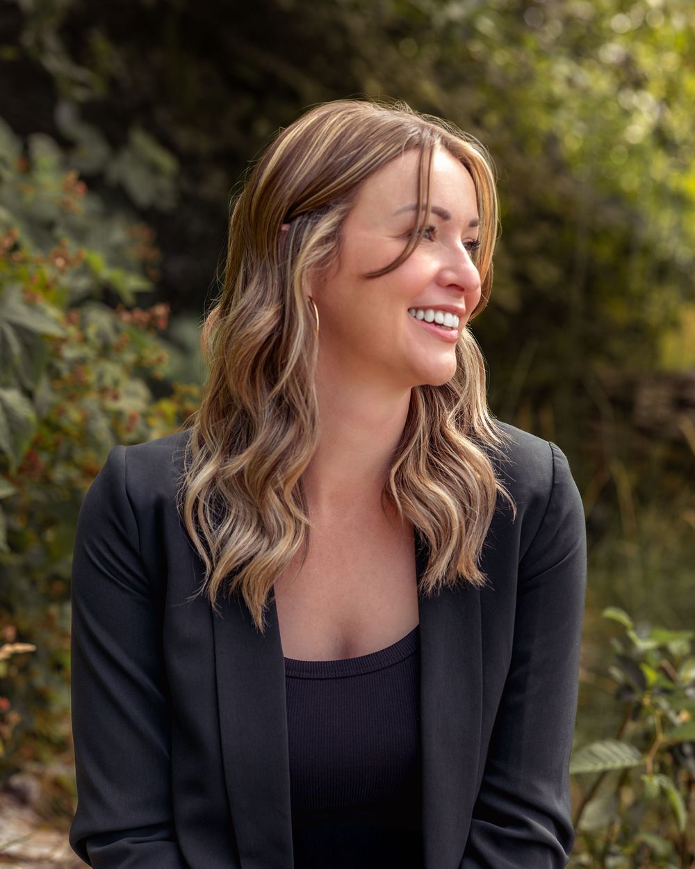 Professional woman in black blazer and wavy hair smiling while looking to the side outdoors.