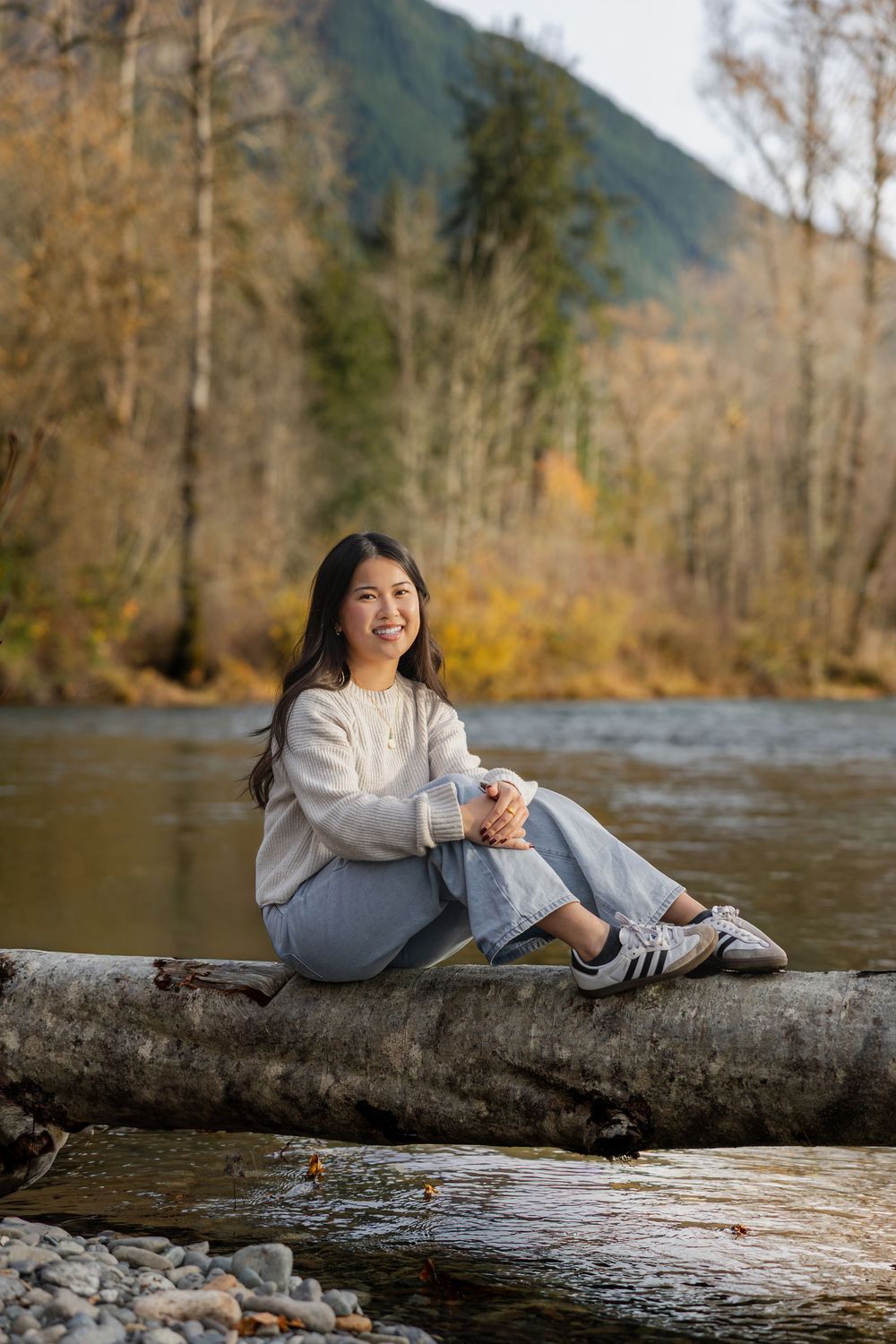 Someone relaxes on a fallen log over a scenic river surrounded by autumn trees and mountains in the background.