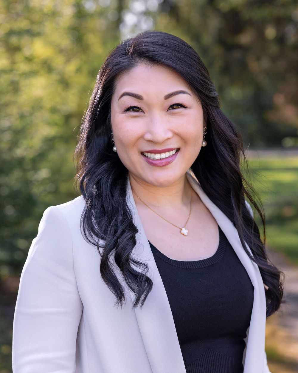 Professional headshot of someone in a white blazer and black top smiling outdoors with blurred greenery in background.