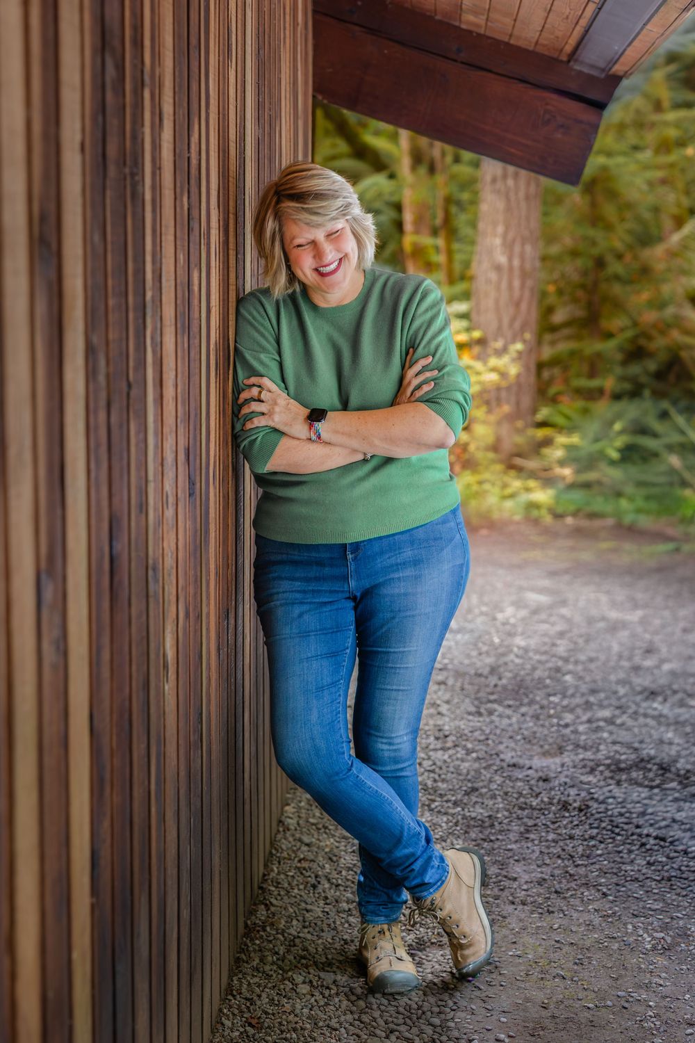 A person wearing a green sweater and blue jeans leans against a wooden post in a forest setting.