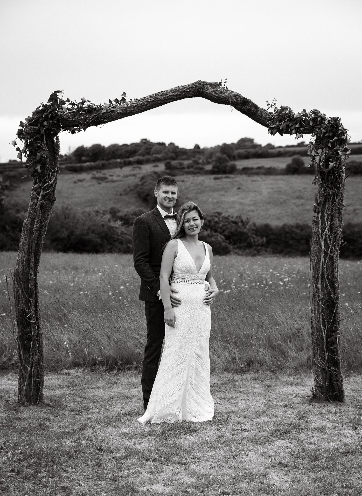 Wedding couple standing under rustic floral arch in countryside setting.