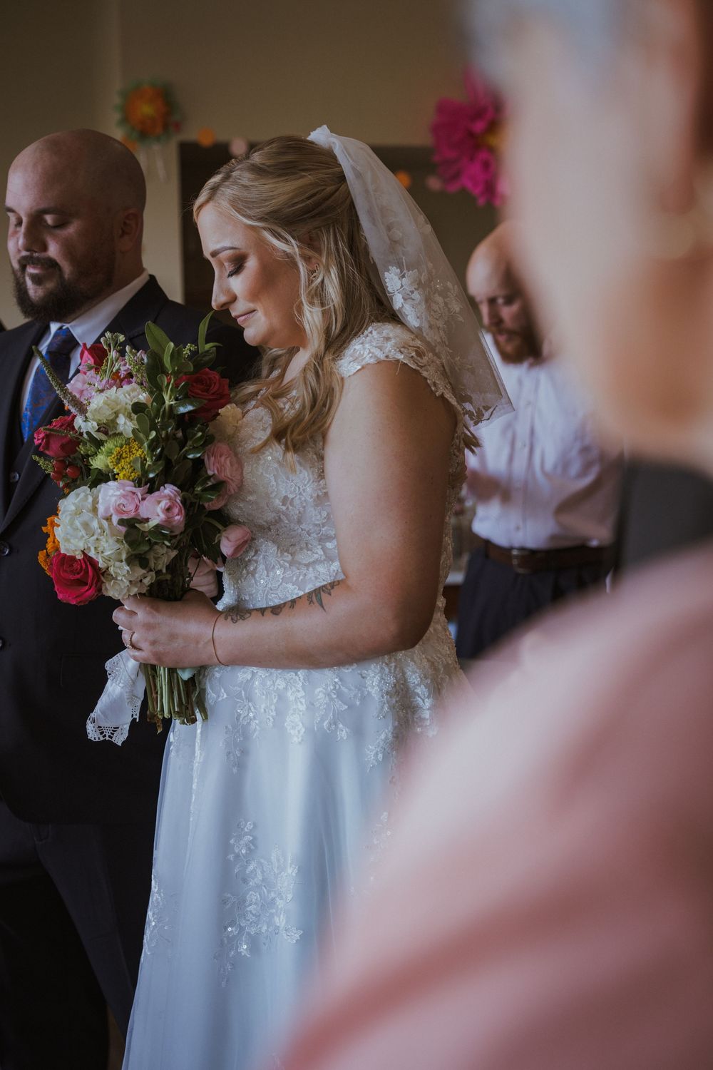 Wedding party gathered indoors with colorful floral bouquets and white lace dress details.