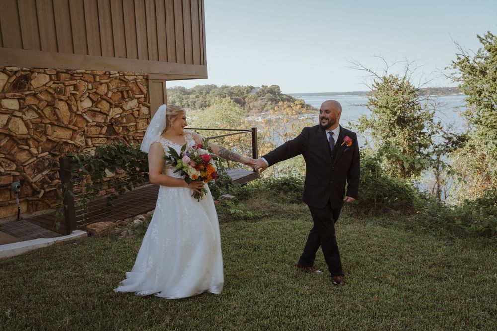 A wedding couple shares a joyful moment on a grassy lawn overlooking a scenic waterfront.
