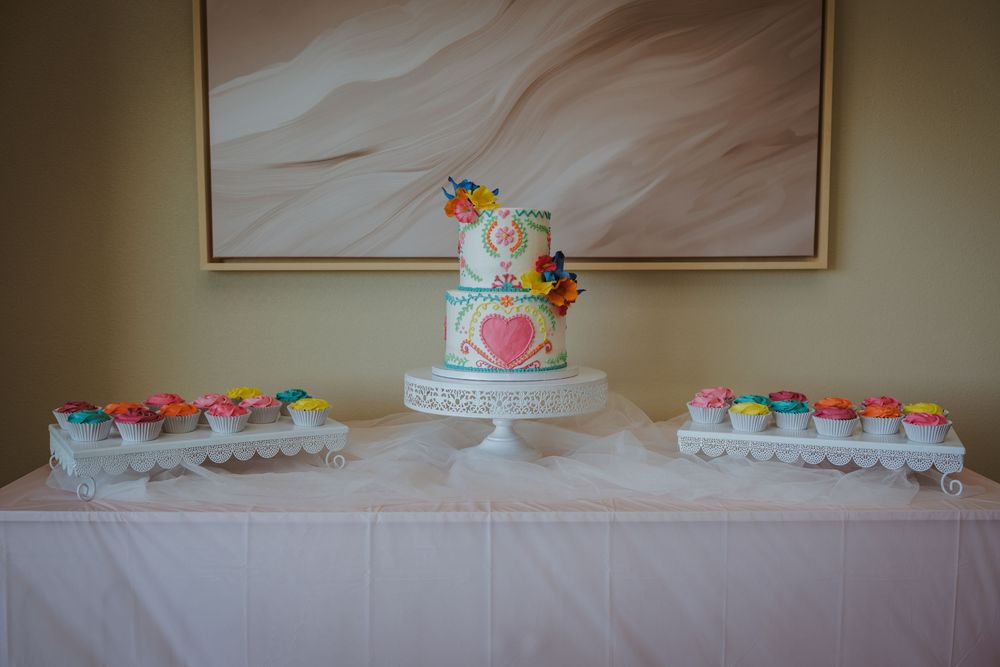 An elegant white wedding cake with colorful floral decorations displayed alongside cupcakes on a dessert table.