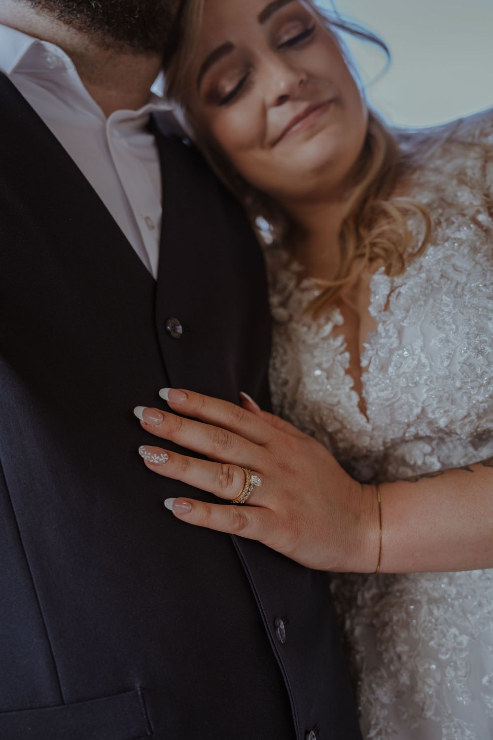Close-up detail of wedding rings and lace dress against dark formal attire.
