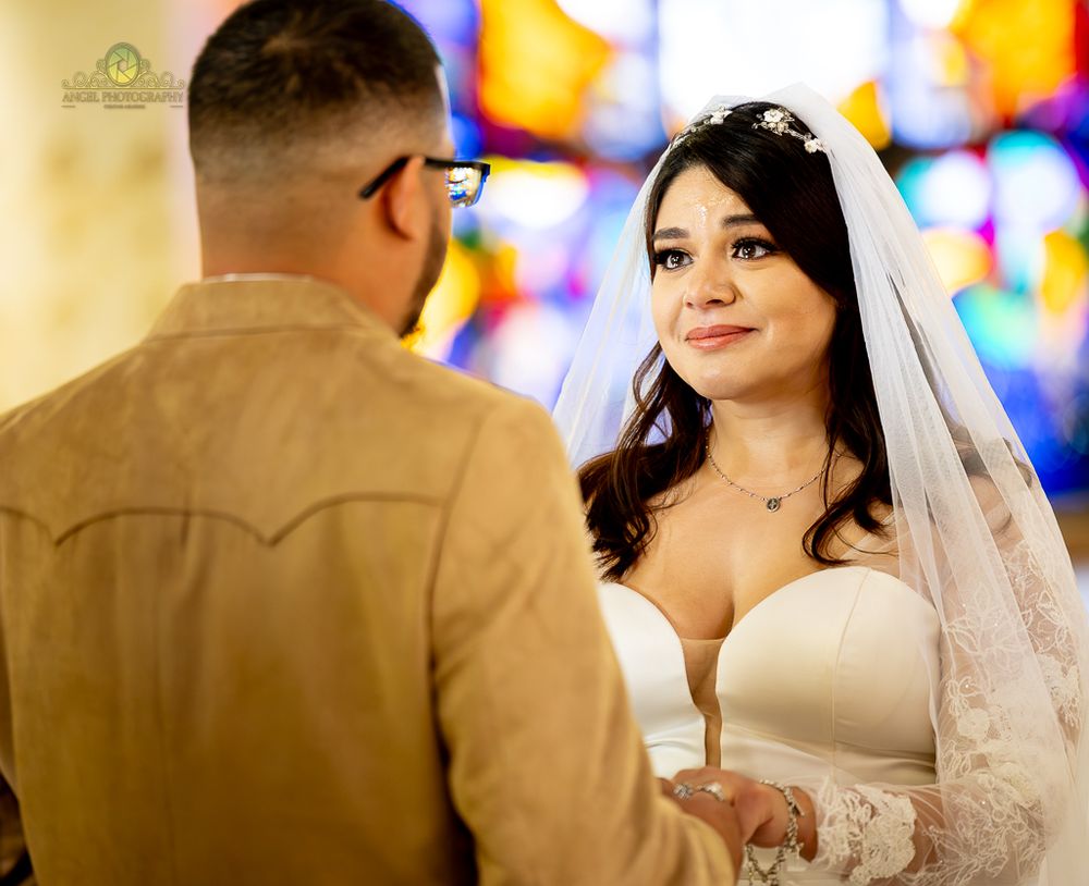 A bride and groom stand together during their wedding ceremony with colorful stained glass windows in the background.