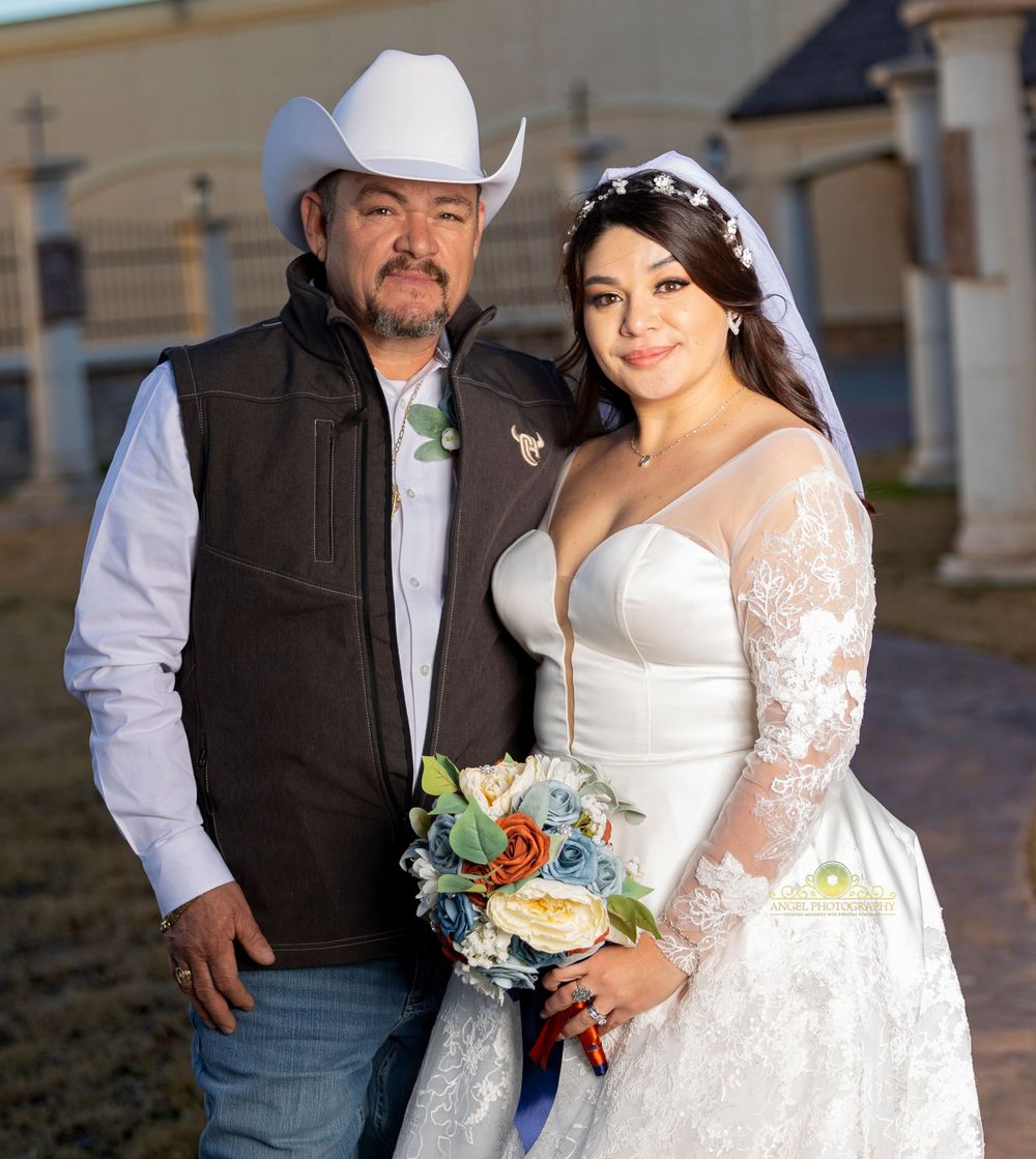 A couple in western wedding attire poses together with bride holding floral bouquet.