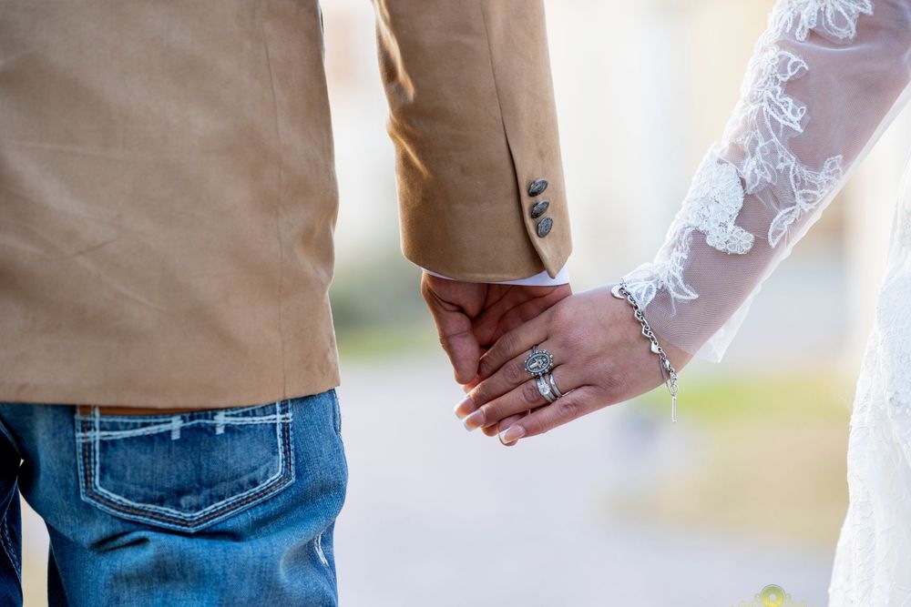 Detail shot of bride and groom holding hands showing lace sleeve and tan jacket.