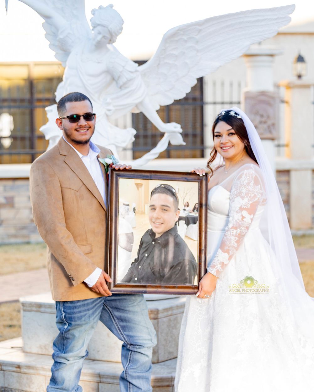 Wedding couple poses with a framed portrait near an angel statue outdoors.