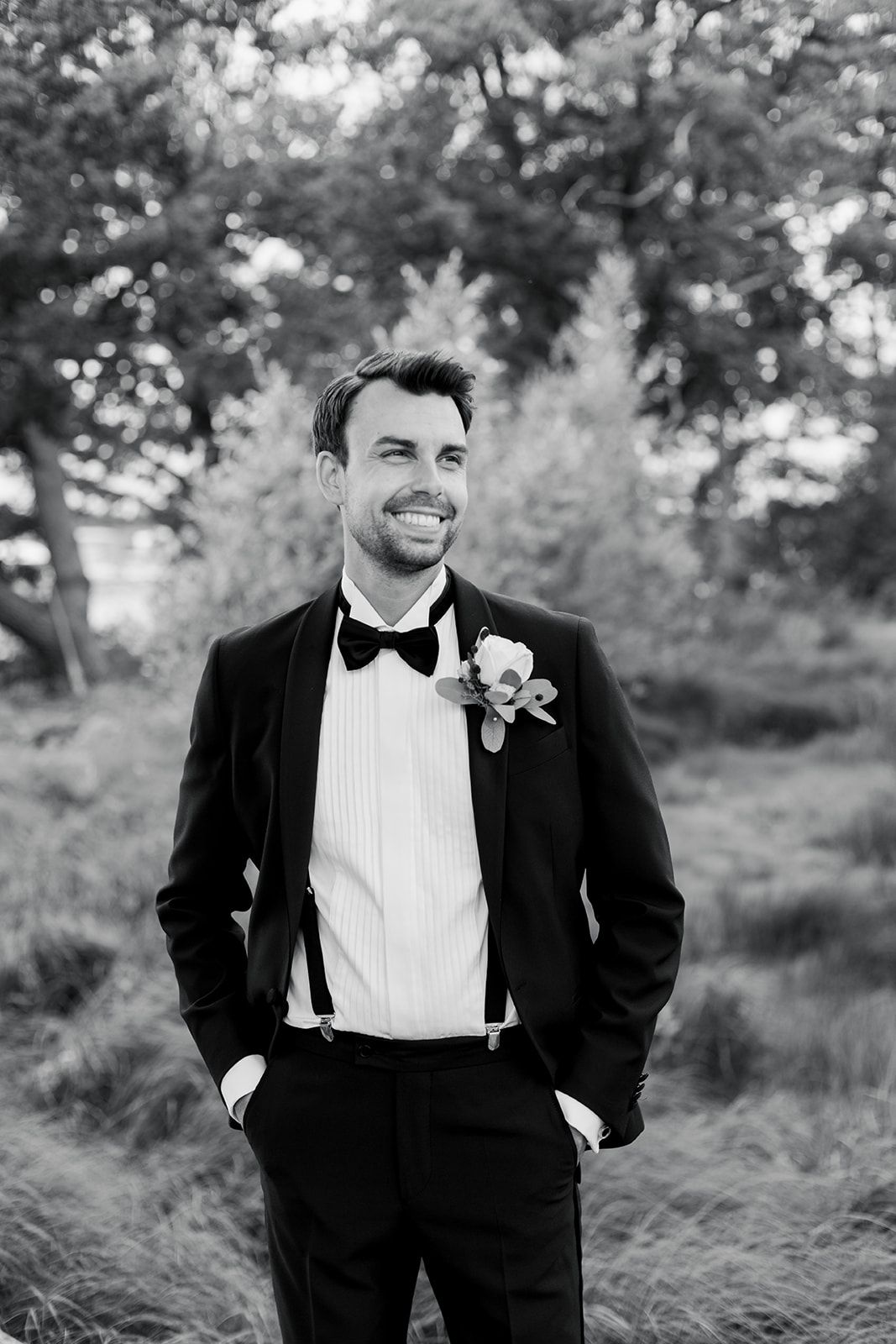 Black and white photo of a groom in classic tuxedo and bow tie smiling outdoors in a natural setting.