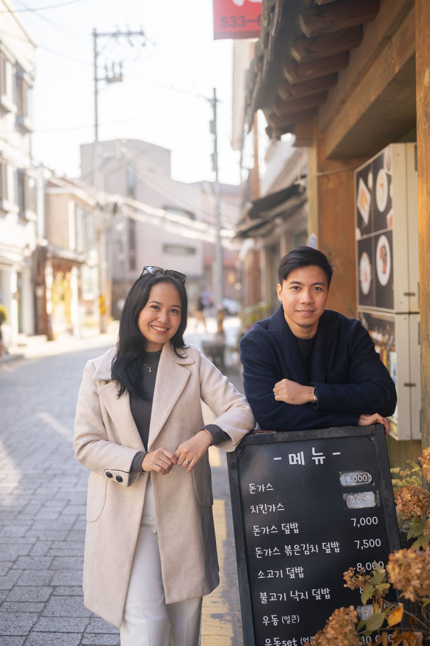 A couple in winter coats poses and smiles along a traditional stone street in Korea.