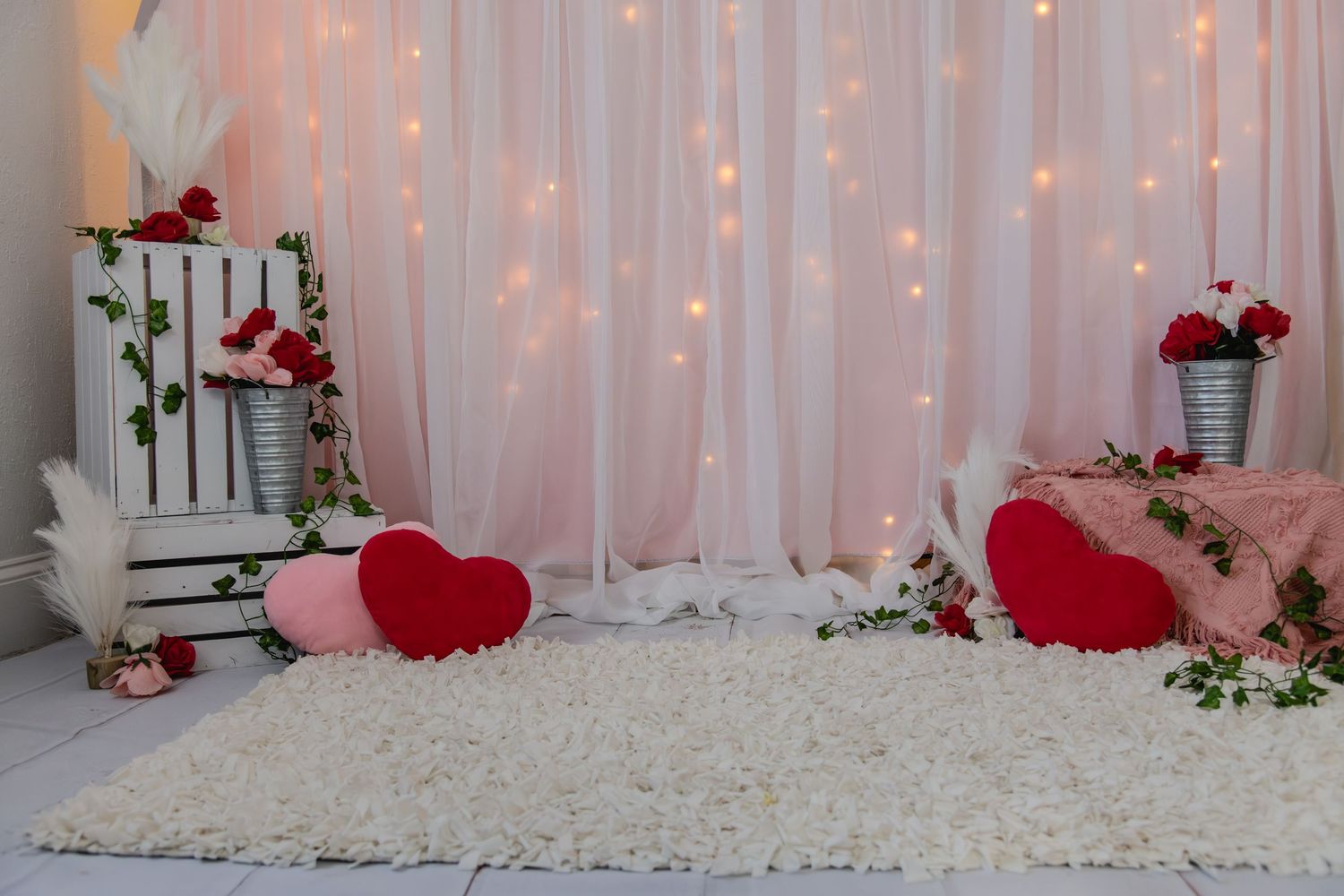 Romantic Valentine's day photo setup with white curtains, string lights, red hearts, and roses on white furry carpet.