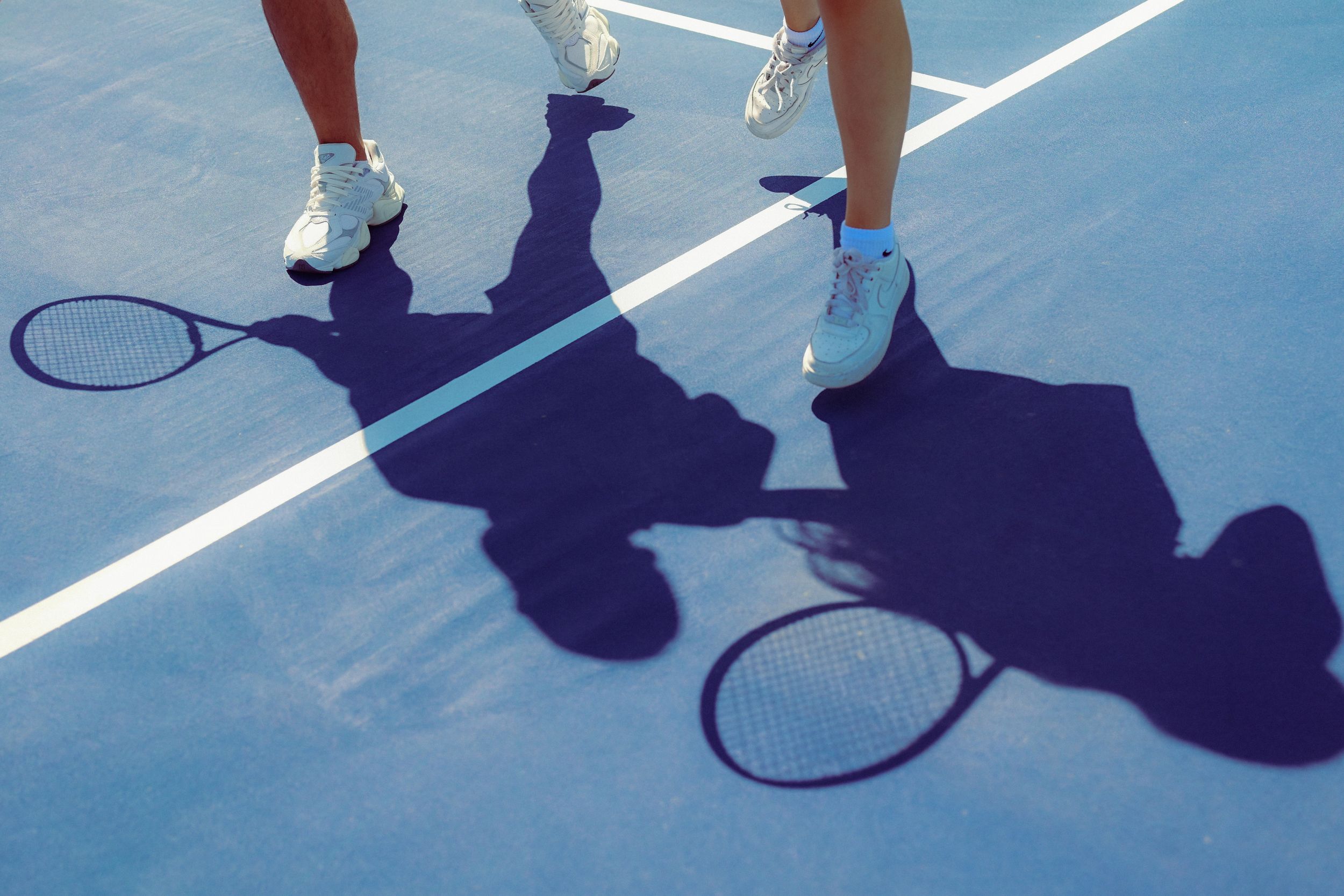 Tennis players casting long shadows on a blue court during a match.