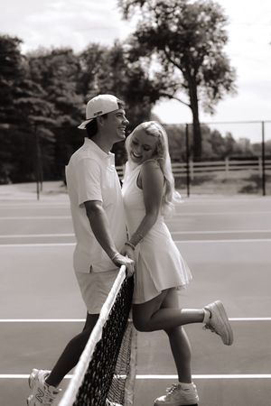 A romantic couple in tennis outfits share playful moments on an outdoor court in black and white.