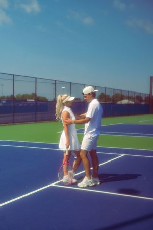 A couple plays tennis together on a bright blue outdoor court on a sunny day.