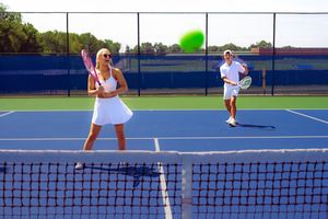 Tennis players compete on blue courts during an outdoor match with nets visible in the foreground.