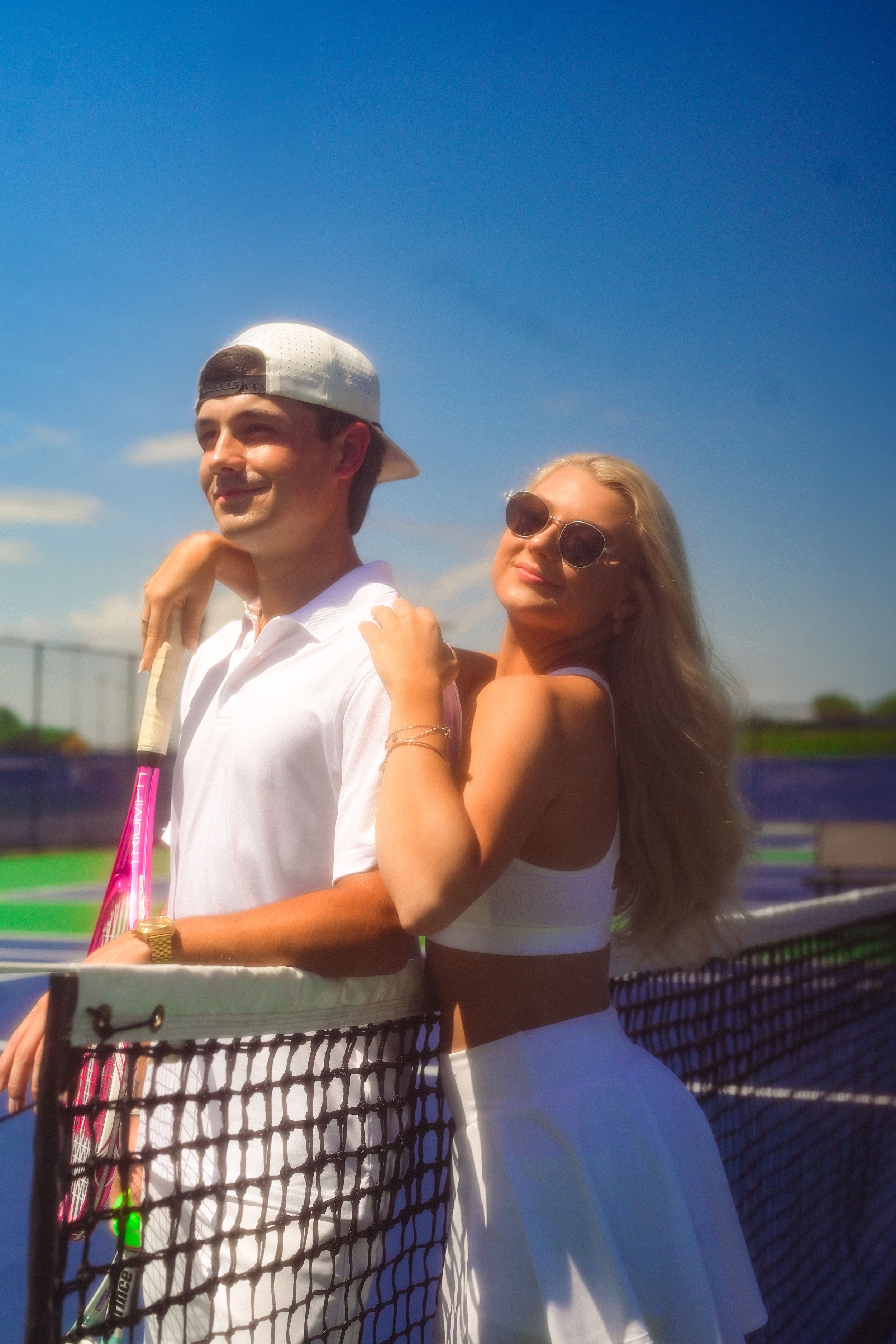 A couple in white tennis attire embraces on a sunny tennis court with blue skies in the background.