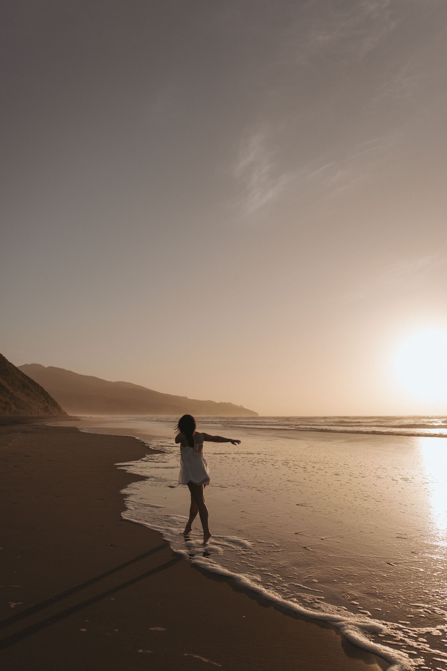 Silhouette of woman in white dress with arms outstretched on sandy beach at golden sunset in New Zealand.