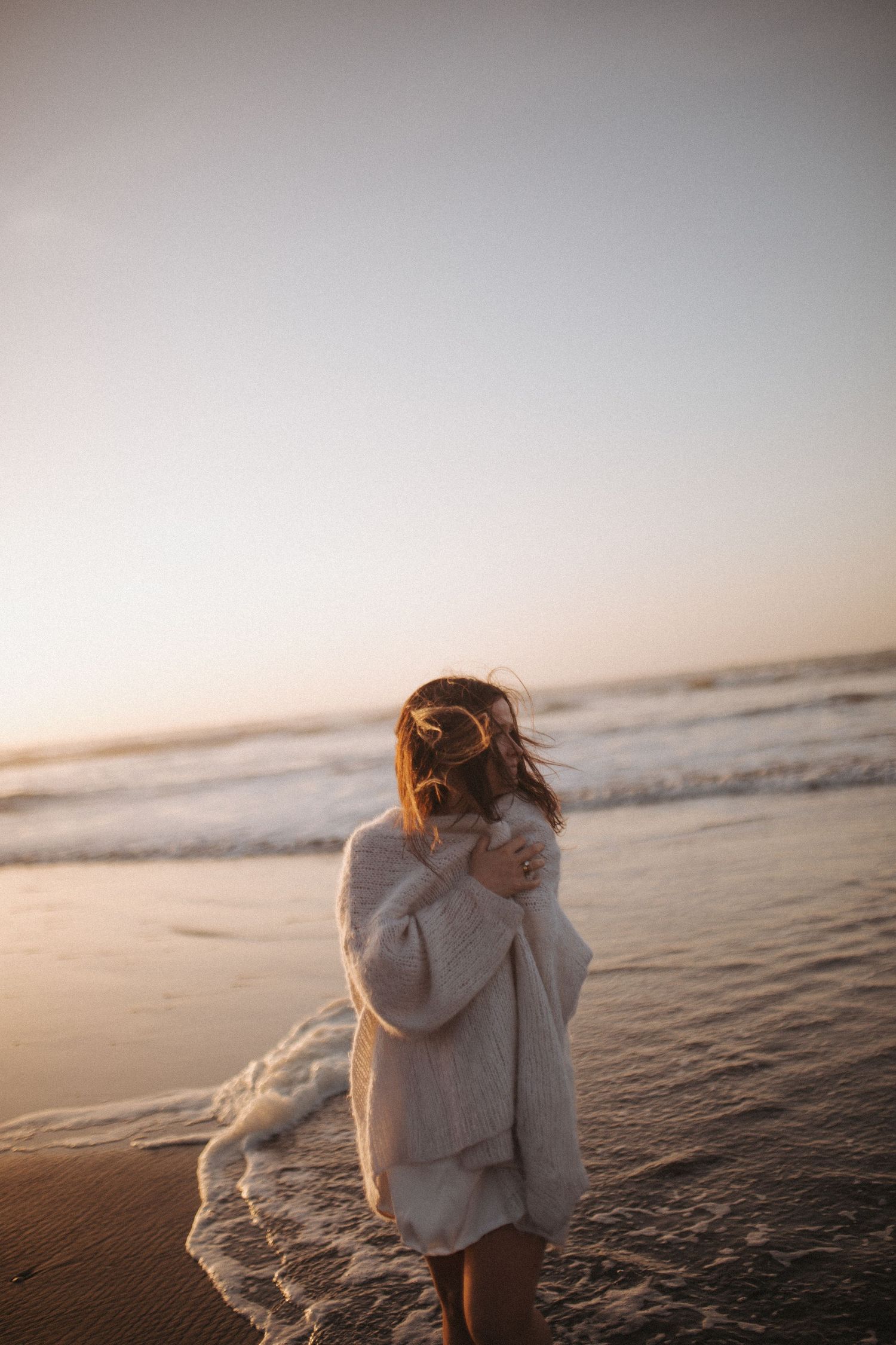 Woman in white fuzzy sweater standing at ocean's edge during golden sunset with waves washing ashore in New Zealand.