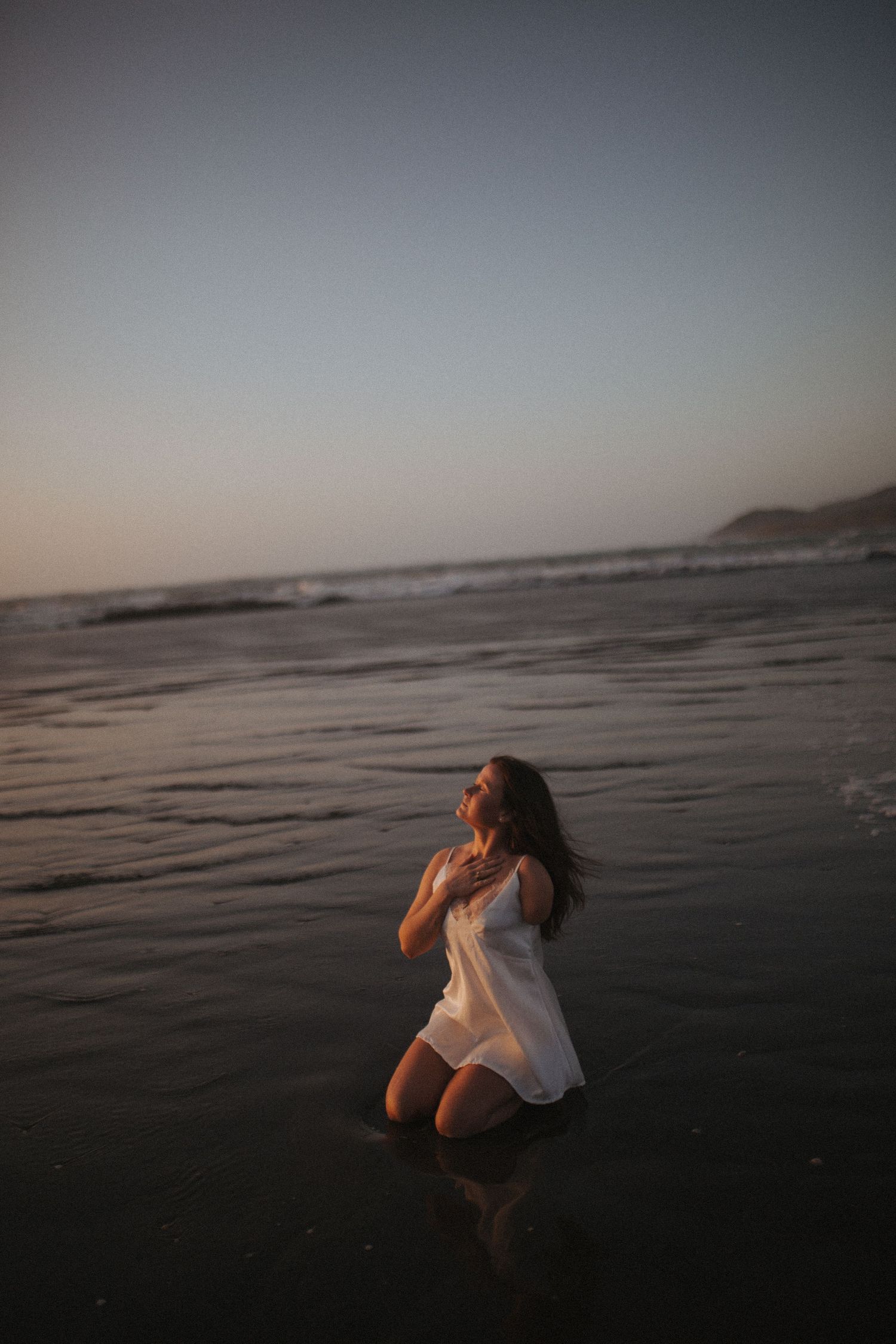 Woman in white dress kneeling in shallow ocean water during a serene beach sunset in New Zealand.