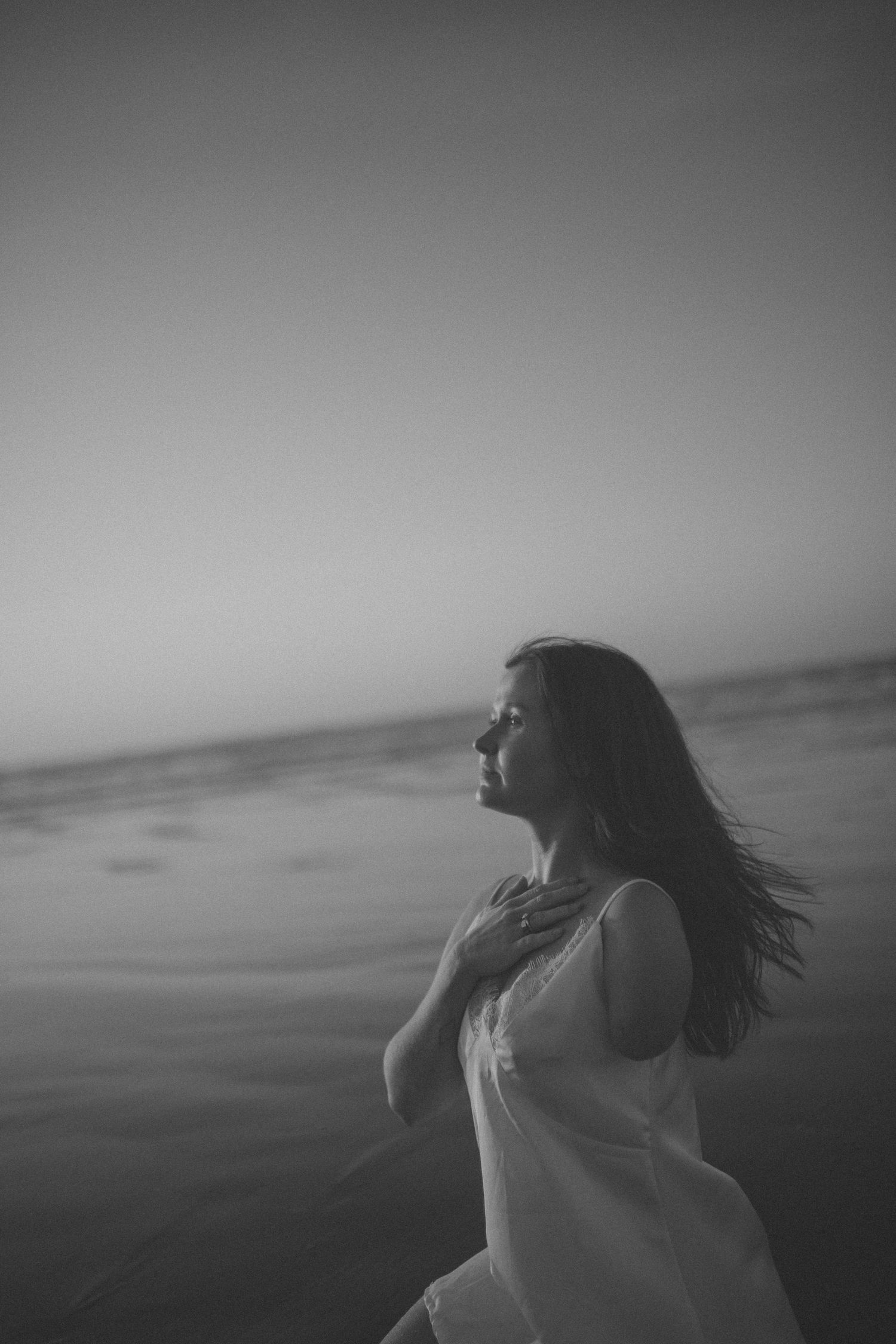 Black and white portrait of woman in white dress with wind-blown hair standing by the sea in New Zealand.