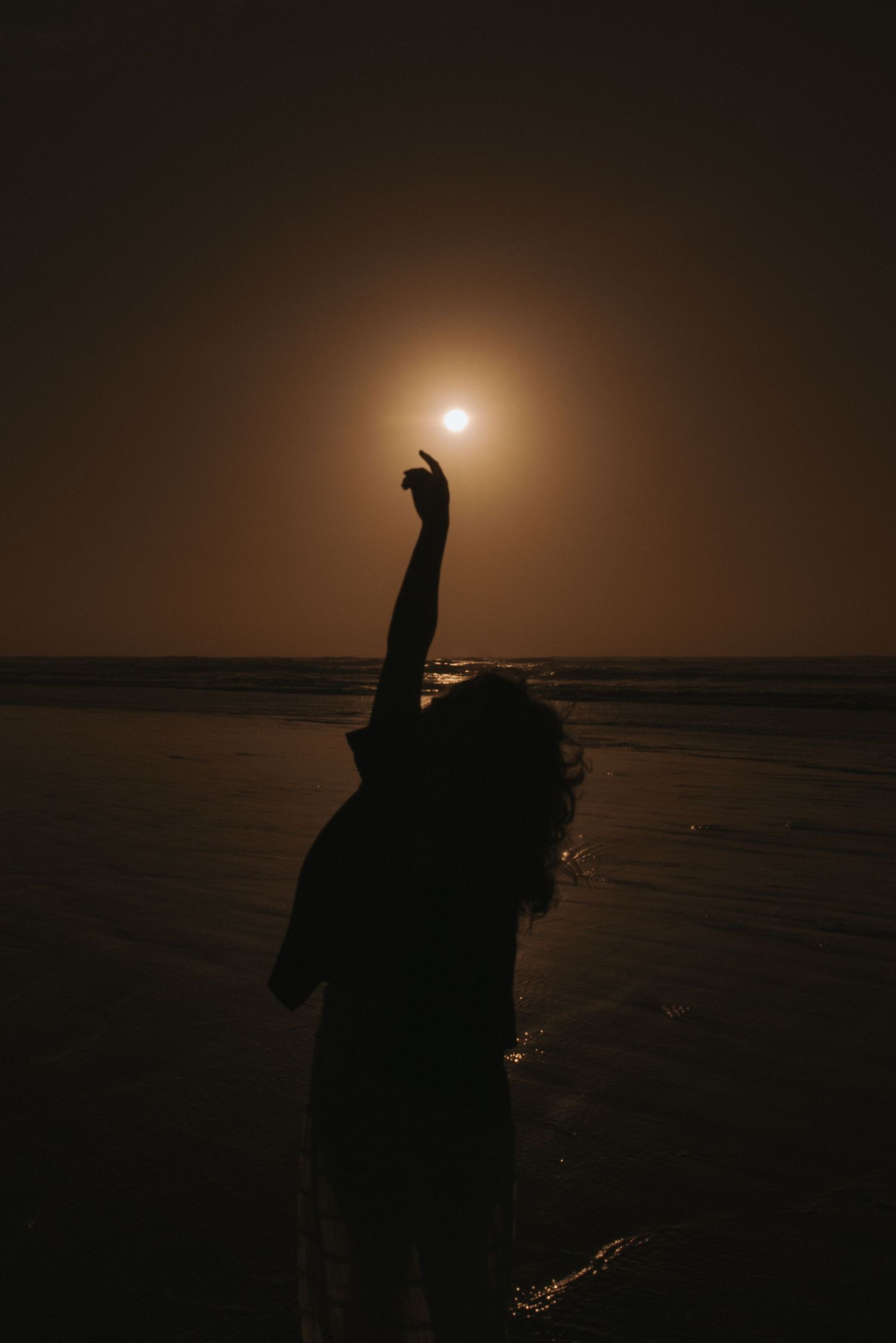 Silhouette of a woman reaching toward the glowing moon on a dark beach at night in New Zealand.