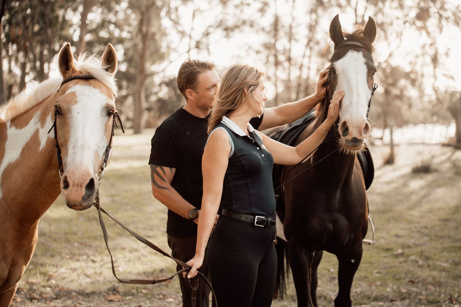 A couple in black attire stands with two horses in a sunlit wooded setting.