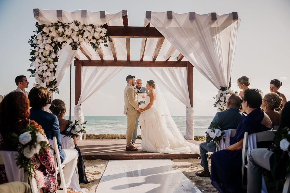 Intimate beach wedding ceremony under wooden pergola decorated with white flowers and fabric. El Dorado Royale Wedding Photographer