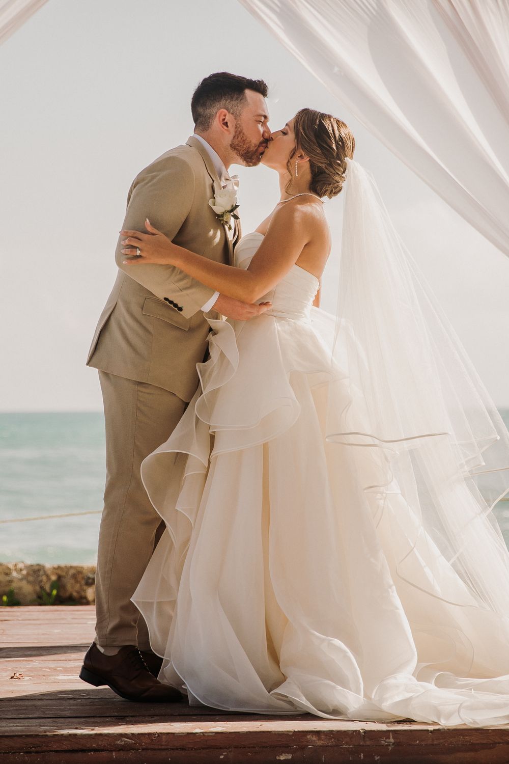 Romantic beach wedding couple shares intimate kisses under flowing white curtains by the ocean. El Dorado Royale Wedding Photographer