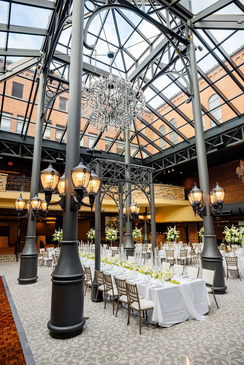Elegant wedding reception setup under a glass atrium with crystal chandelier and vintage-style lamp posts.