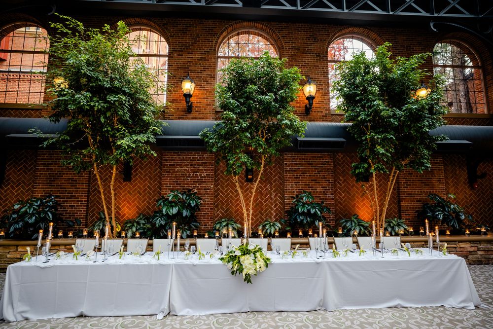 Long white banquet table decorated with greenery against a brick wall with illuminated arched windows.