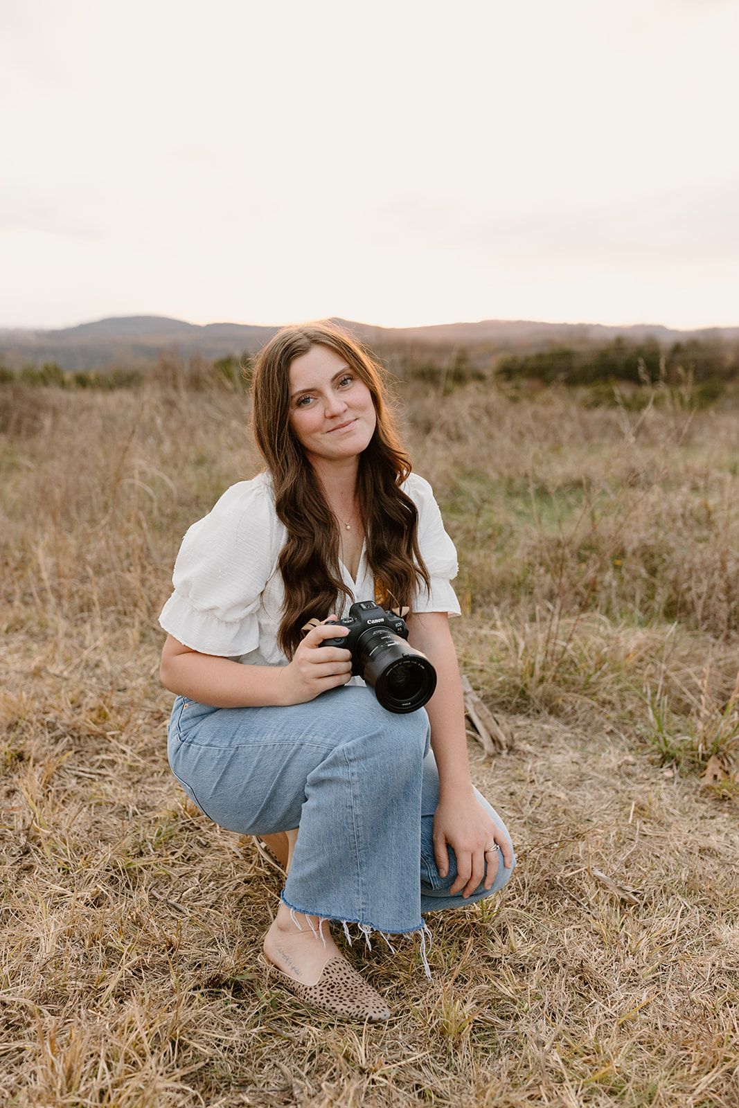 Photographer in white blouse and jeans crouches in dry grass field with camera while smiling at sunset.