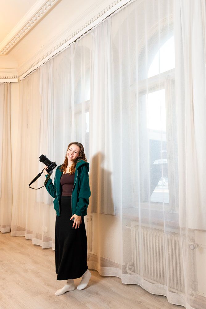 Valokuvaaja Anniina Virtanen, Turku, pitämässä kameraa kädessä ja hymyilemässä lämpimästi kuvaajan suuntaan valokuvausstudiolla.