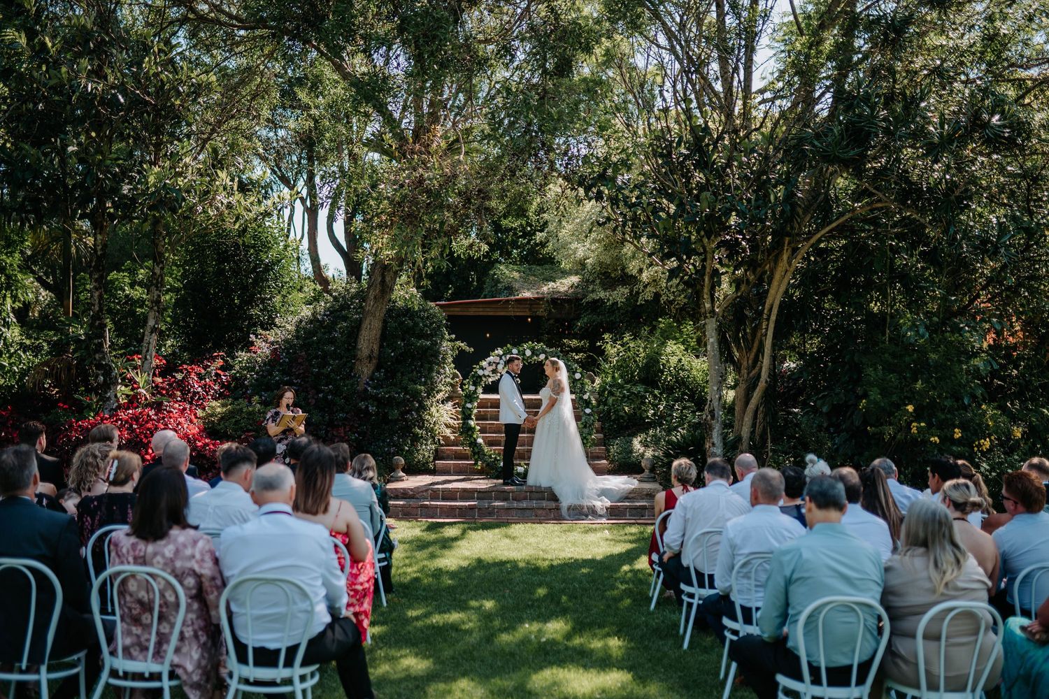 A beautiful outdoor wedding ceremony setup with white chairs arranged in rows under tall trees on a sunny day.